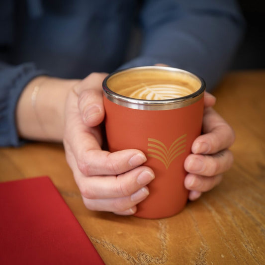 Person holding a red insulated mug with a drink on a wooden surface